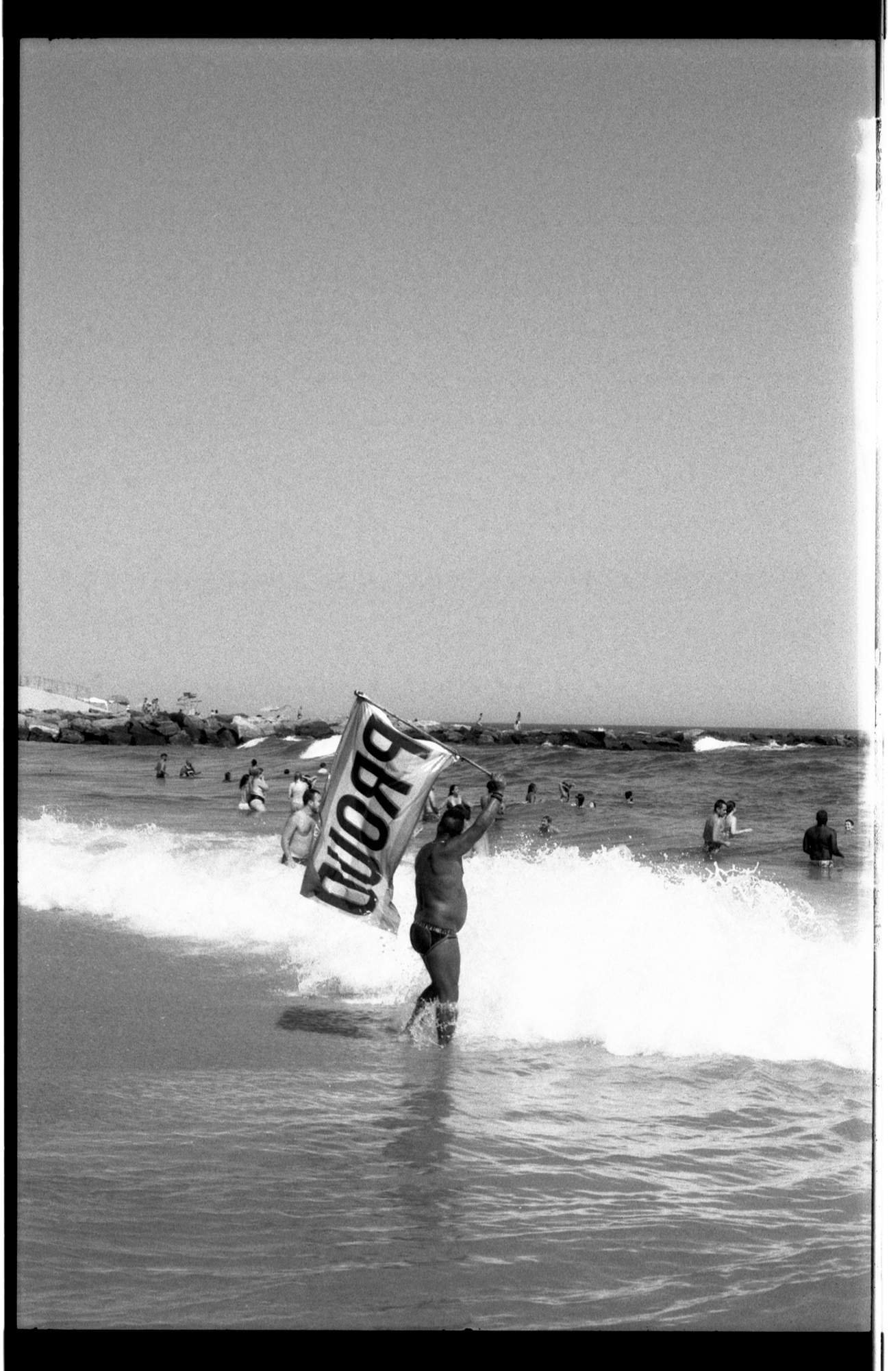 A speedo-clad bather wades into the surf at Riis Beach, carrying a large flag displaying the word "PROUD."