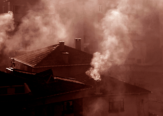 Photograph. Smoke billows out from homes in a crowded urban neighborhood.