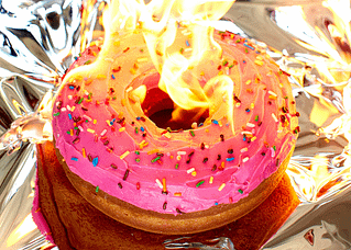 A donut with pink frosting and sprinkles is on fire. Tinfoil under and behind the donut increases the drama of the photo.