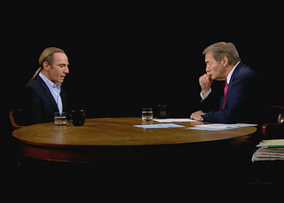 Screen capture from John Galliano’s interview on Charlie Rose in 2013. Seated at large round table in formal wear, Galliano and Rose are pictured in conversation against a black backdrop.