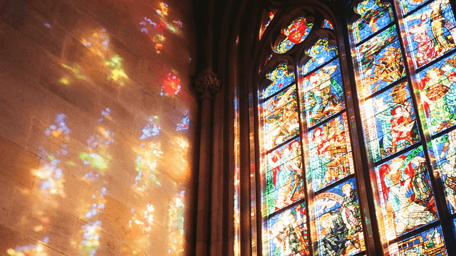 Sunlight streams through stained-glass windows in a church, casting colorful patterns against an adjacent stone wall.