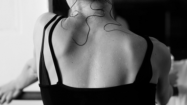 Close-up photograph of a dancer’s back. She is wearing a black leotard. Her back is visibly sweaty, with strands of hair pasted down on her skin.