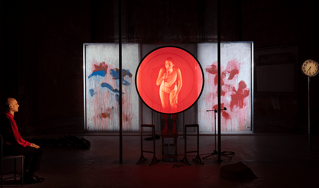 Production photo from the Italian stage adaptation of Katharina Volckmer’s The Appointment. The lead actress is standing up on a chair. She is washed in red by a large transparent disc of light between her and the audience. In the background is a paint splattered wall. Off to the left side, the doctor character is seated with his eyes closed.