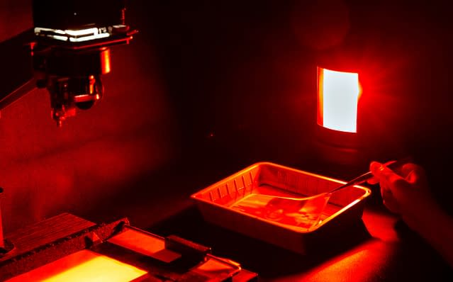 Inside a darkroom, a red light illuminates a hand using tweezers to gingerly lift a print out of a water bath.