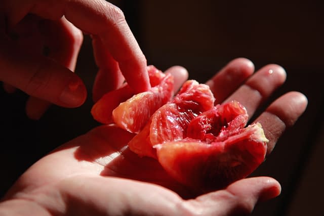 Five wedges of blood orange rest on an open palm. The subject's other hand is suggestively touching two of the wedges.