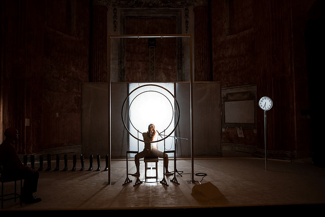 Production photo from the Italian stage adaptation of Katharina Volckmer’s The Appointment. The lead actress is seated facing away from the audience. She gazes at a bright light at the back of the stage. Off to the left side, the doctor character is seated with his eyes closed.