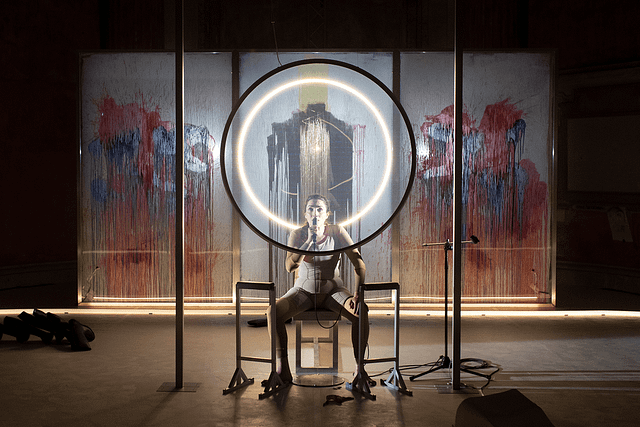 Production photo from the Italian stage adaptation of Katharina Volckmer’s The Appointment. The lead actress is seated and gazing forward at the audience. Ahead and behind are giant ring lights illuminating her face. In the background is a paint splattered wall.