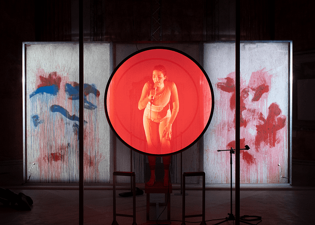 Production photo from the Italian stage adaptation of Katharina Volckmer’s The Appointment. The lead actress is standing up on a chair. She is washed in red by a large transparent disc of light between her and the audience. In the background is a paint splattered wall. Off to the left side, the doctor character is seated with his eyes closed.