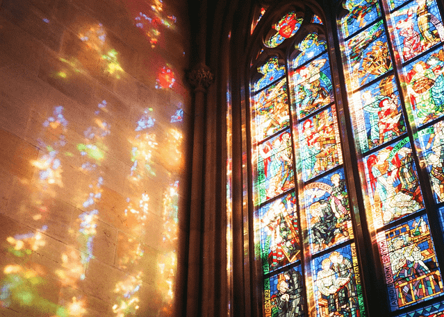 Sunlight streams through stained-glass windows in a church, casting colorful patterns against an adjacent stone wall.