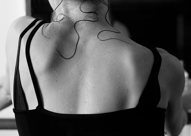 Close-up photograph of a dancer’s back. She is wearing a black leotard. Her back is visibly sweaty, with strands of hair pasted down on her skin.