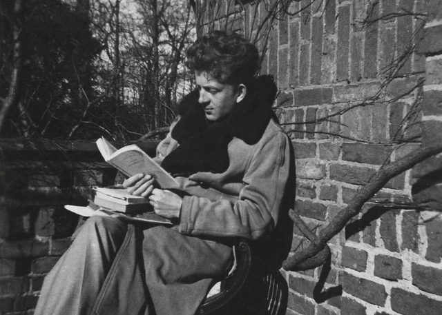 Photograph of the Dutch poet Hans Lodeizen (1924-1950). He is seated outdoors in a heavy coat, reading while also resting several other books in his lap. The photograph was taken in his hometown of Wassenaar.