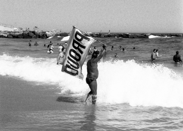 A speedo-clad bather wades into the surf at Riis Beach, carrying a large flag displaying the word 