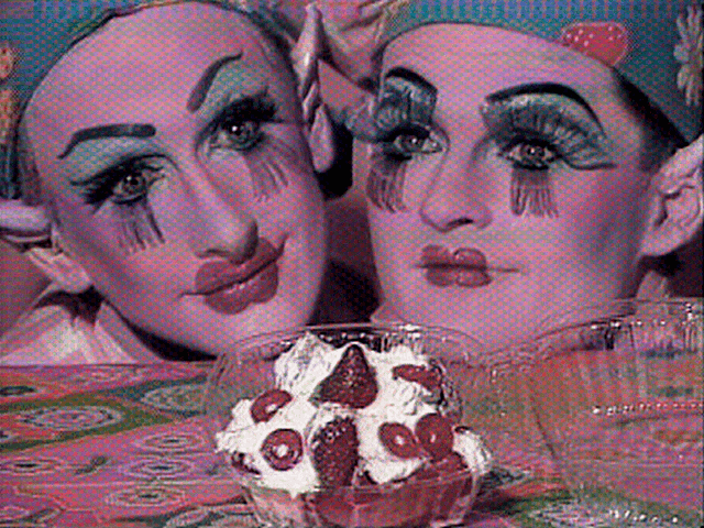 Two pink faces, painted like clowns with blue eyeshadow and caps, resting on a table and staring at a punnet of strawberries in cream, in a still taken from Tom Rubnitz's 1989 film strawberry shortcut