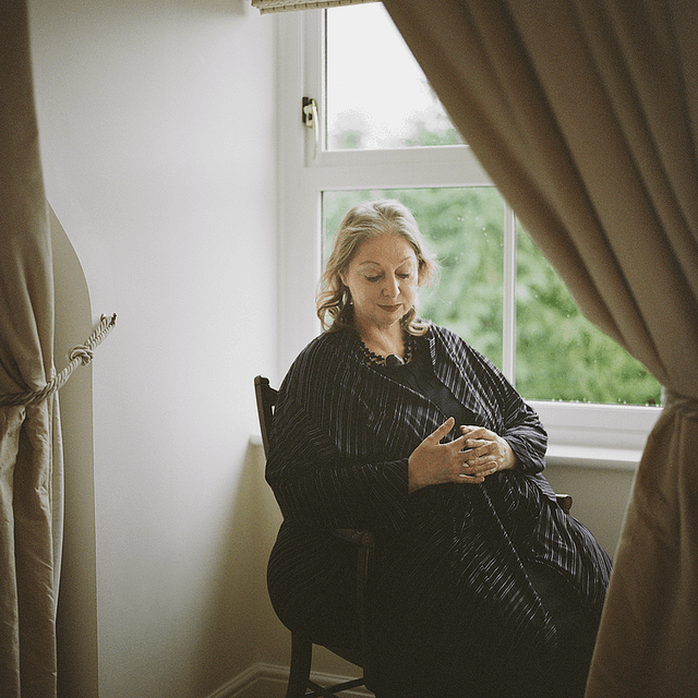 Portrait of Hilary Mantel in 2020. The author is seated by a window. She laces her hand in front of her stomach and is gazing downward. The light is soft.